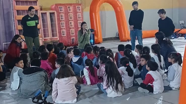 En la imágen se visualiza un grupo de niños sentados escuchando a tres adultos que les brindan una charla sobre seguridad vial. Alrededor de ellos hay una pista inflable y un karting a pedal.