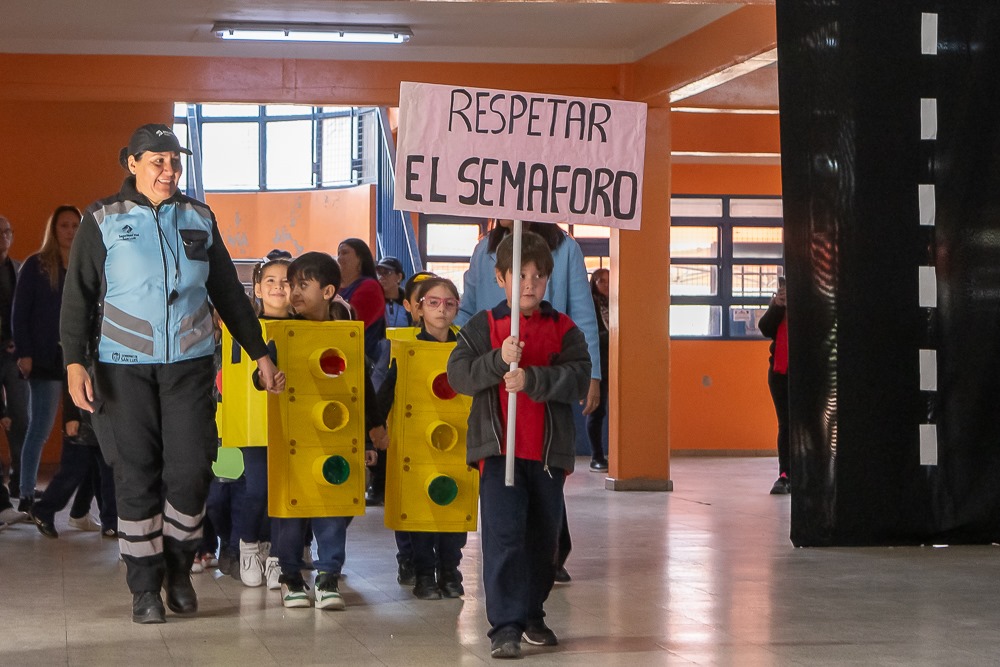 En la imágen se ven niños y niñas en edad escolar disfrazados de semáforos. Van caminando de la mano de una Agente de Tránsito. Es un desfile. El Primer niño lleva un cartel que dice "Respetar el semáforo"