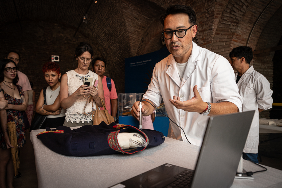 La imagen muestra al conservador del Museo Casa Rosada César Jacobi sosteniendo una lupa digital con su mano derecha. Viste un guardapolvo blanco y lleva gafas negras. Apoya la lupa sobre una chaqueta militar de color azul con detalles rojos y dorados. Mira atento la pantalla de una computadora portátil. Por detrás de él un grupo de cinco personas observa la actividad, una de ellas toma una foto con su teléfono móvil. La escena ocurre en una de las salas del Museo.