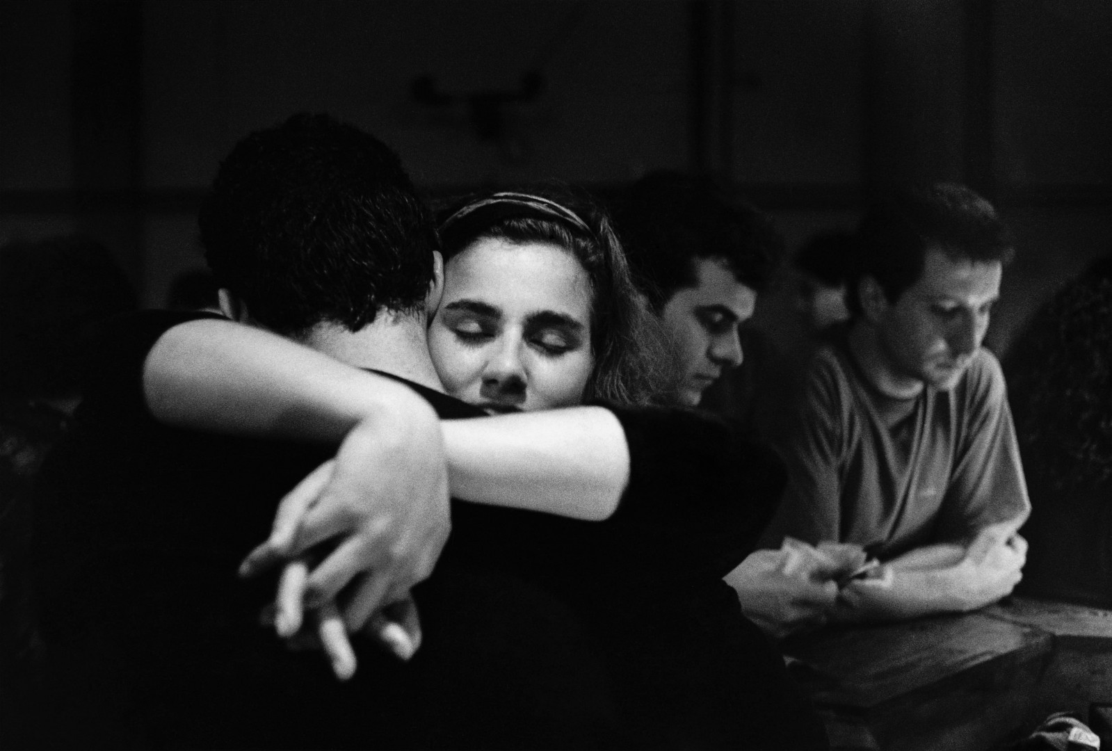 En esta fotografía en blanco y negro de Adriana Lestido titulada "La salsera", se observa a una mujer abrazando. Detras, se observan dos hombres apoyados en una barra. 