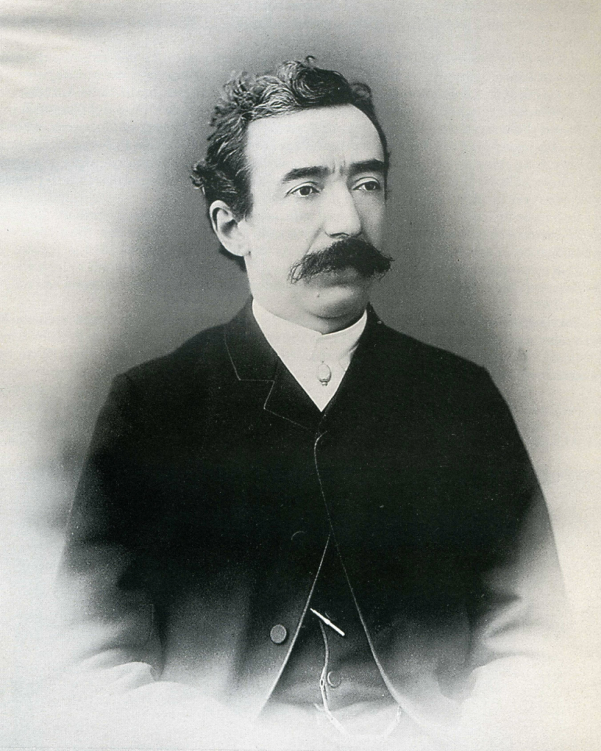 La imagen es una fotografía en blanco y negro de un hombre llamado Francisco Tamburini con bigote y cabello ondulado. Lleva puesto un traje oscuro con una camisa blanca y un cuello alto.