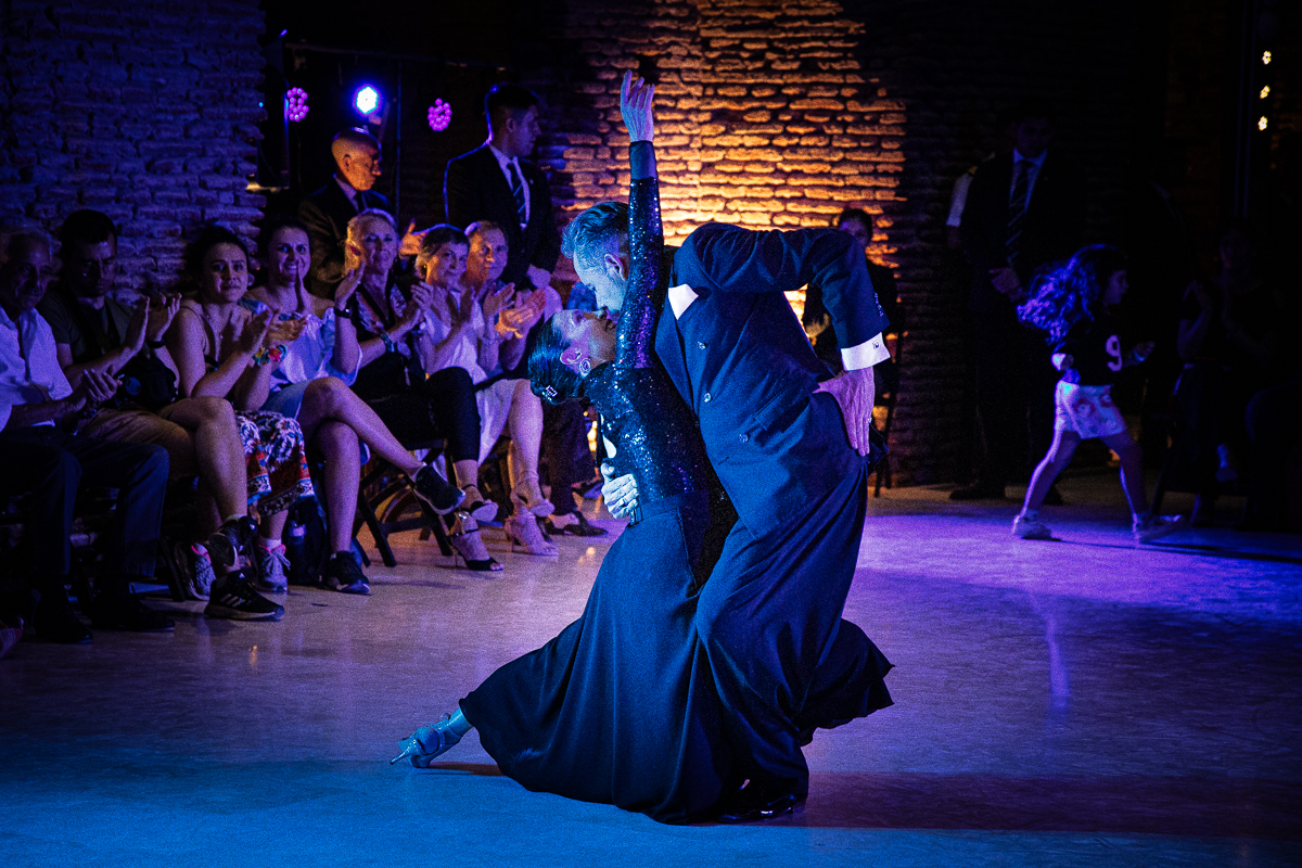 La imagen muestra una pareja bailando tango. Ambos están vestidos de negro y están en una pose elegante y dramática. Al fondo, se puede ver a un grupo de personas sentadas, observando la actuación. La iluminación es tenue, con luces azules y cálidas que resaltan a los bailarines. Al fondo se observa una pared de ladrillos.