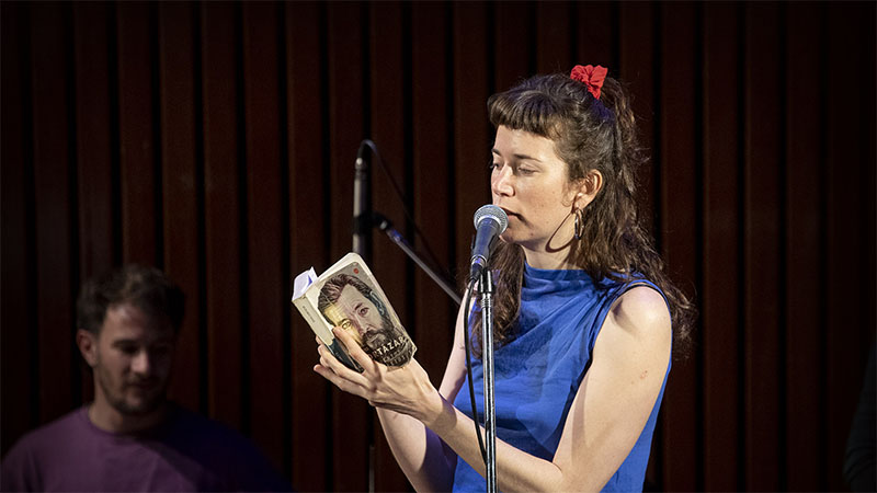 Una mujer está parada frente a un micrófono leyendo un libro de Julio Cortazar al público.