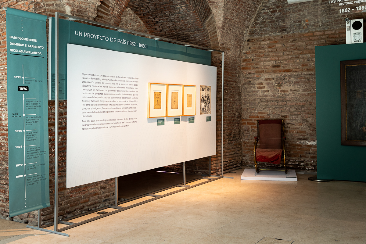 La imagen muestra una exhibición en un museo o galería. Hay un panel informativo grande en la pared con texto y algunas imágenes enmarcadas. El texto del panel dice "Un proyecto de país (1862-1880)". A la izquierda del panel, hay una línea de tiempo vertical con varios eventos marcados. La pared de fondo está hecha de ladrillos y el suelo es de color claro. También hay una silla mecedora.