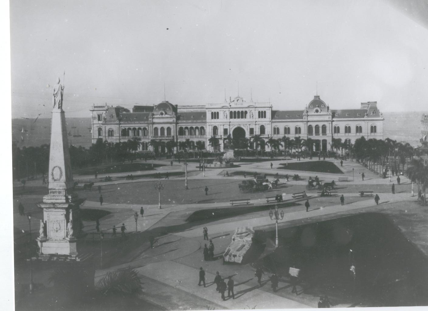 La imagen muestra una fotografía en blanco y negro de la Plaza de Mayo. A la izquierda y en primer plano se observa la Pirámide de Mayo. En el fondo se puede ver la Casa Rosada, edificio de arquitectura clásica. En la plaza se ven los caminos y algunas personas recorriendo la plaza.