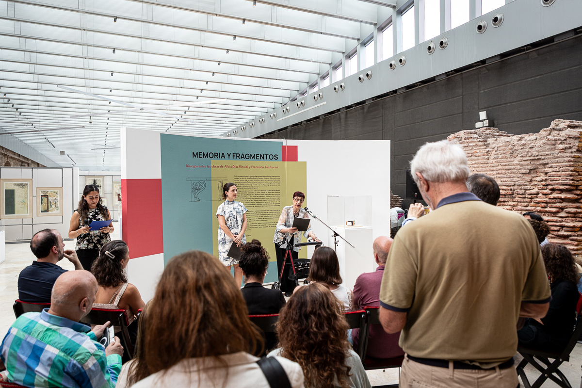 La imagen muestra un evento en un espacio interior, probablemente un museo, donde varias personas están sentadas y prestando atención a una presentación. Al fondo hay un panel grande con texto y el título "Memoria y fragmentos". Dos personas están de pie frente al panel, una de ellas parece estar hablando. 