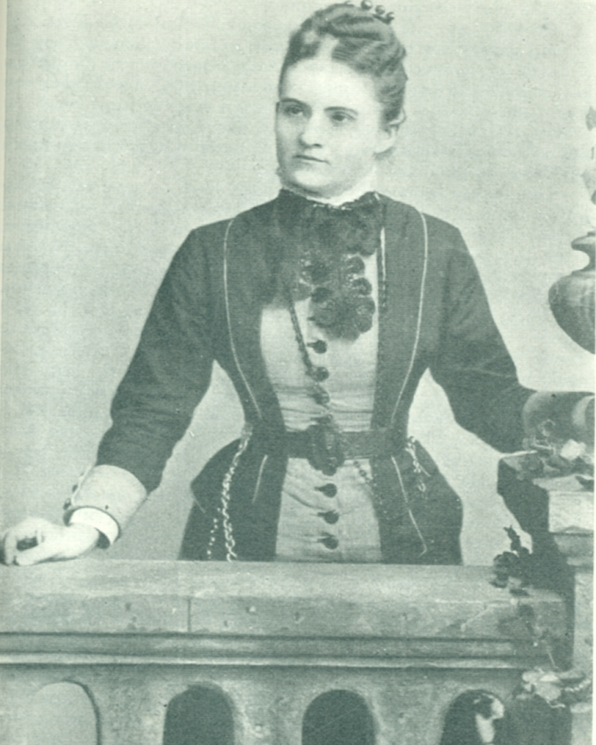 Sara Chamberlain de Eccleston La imagen muestra una fotografía antigua en blanco y negro de una mujer de pie. Está vestida con un vestido de estilo victoriano, que tiene detalles oscuros y claros, con lazo en el cuello. Su cabello está recogido. Ella está apoyada sobre una barandilla o un mueble, su mano derecha descansando sobre él.