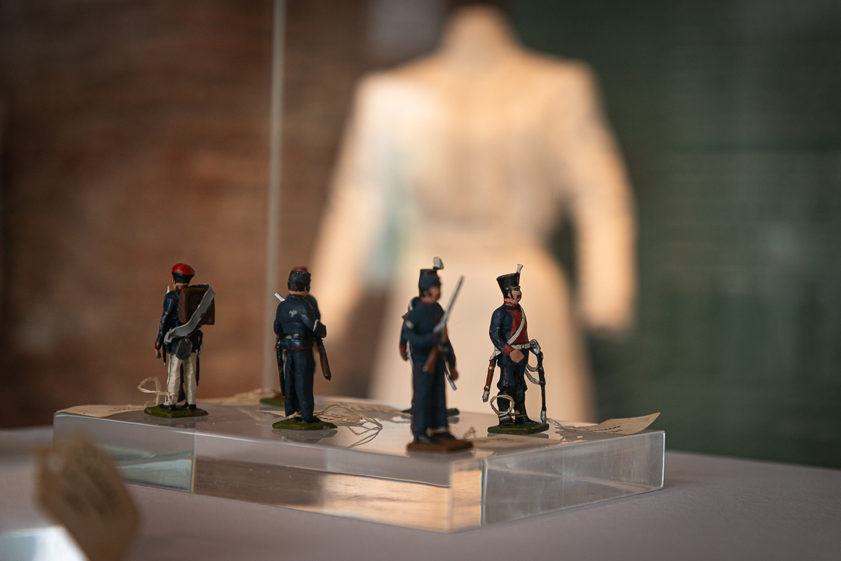 La imagen muestra varias figuras en miniatura de soldados, colocadas sobre la superficie transparente. Las figuras están alineadas en fila y parecen estar vestidas con uniformes militares de época. La imagen está tomada desde un lateral, mostrando a los soldados de perfil. El fondo es borroso, lo que resalta más las figuras en primer plano.