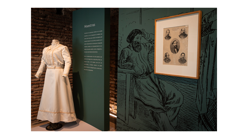 La imagen muestra una exhibición en un museo. En la parte superior hay un maniquí vestido con un traje femenino blanco antiguo. Al lado del maniquí, hay un panel verde con texto cuyo título es "Maestras". Hacia la derecha hay una ilustración en la pared que representa a un gaucho con barba. Sobre la pared verde hay un cuadro enmarcado con varias fotografías en blanco y negro de personas, posiblemente figuras históricas relacionadas al tema de la exhibición