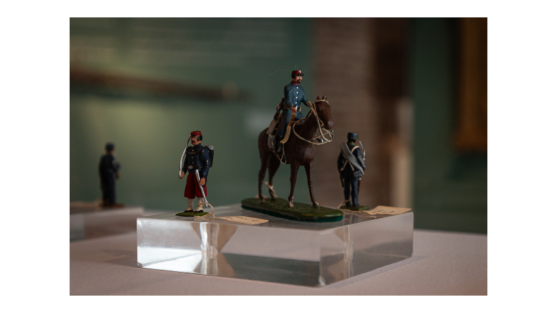 La imagen muestra varias figuras en miniatura de soldados. Visten uniformes históricos. Las figuras están colocadas sobre una base transparente. El fondo está desenfocado.