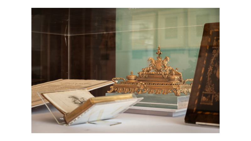 La imagen muestra una exhibición en el museo. Al interior de una vitrina hay un objeto dorado en el centro, un tintero de bronce con detalles intrincados y formas curvas. A la izquierda hay un libro abierto, mostrando una página que tiene el retrato de Sarmiento. En la parte inferior se ve la esquina de una carpeta de madera.