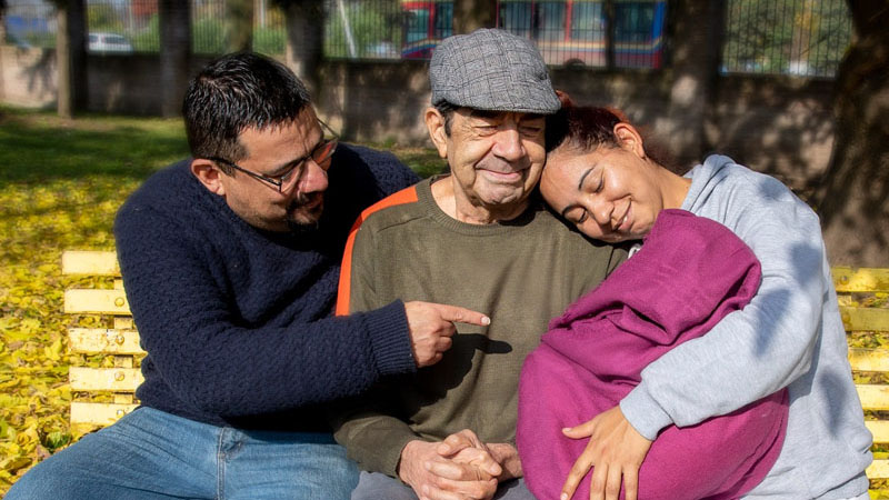 Adulto mayor junto a dos personas sentados en el banco de una plaza.